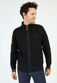 Veste en polaire noire avec col montant, fermeture éclair intégrale sur le devant et tirette orange. Présente un logo brodé et des accents rayés aux poignets.
