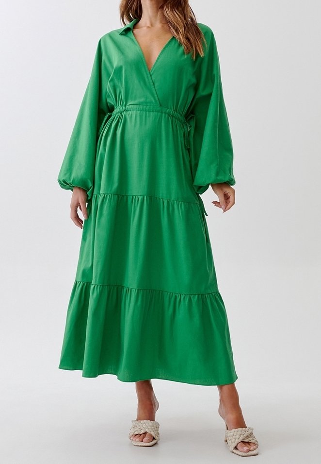Femme portant une longue robe verte à étages avec des manches ballon et des sandales beiges tressées à talons, debout devant un fond uni.