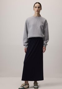 Donna in piedi che indossa una felpa grigia, una lunga gonna nera e scarpe da ginnastica verdi e bianche su uno sfondo chiaro e uniforme.