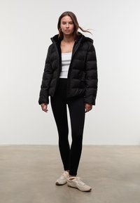 Schwarze Daunenjacke mit strukturierter Oberfläche, Kapuze und Seitentaschen, getragen über einem weißen Oberteil und kombiniert mit schwarzen Leggings sowie beigefarbenen Sneakers.