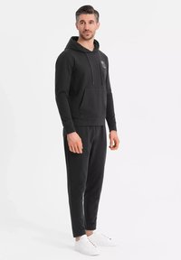 Svart huvtröja med en framficka och snörning, kombinerad med matchande joggingbyxor och vita sneakers. Slät textur med minimal märkning.