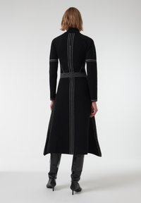 Zwarte lange jas met een hoge kraag, voorzien van witte stiklijnen langs de naden, een getailleerde taille en een uitlopende silhouet. Gestyled met kniehoge laarzen.