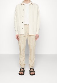 Giacca button-up beige chiaro con colletto e tasche, abbinata a pantaloni cargo beige chiaro e sandali neri con doppia fibbia.