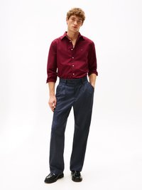 Rode katoenen shirt met knoopsluiting, gecombineerd met marineblauwe geblokte broek en zwarte nette schoenen. Model staat met de handen in de zakken.