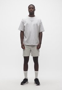 Άνδρας όρθιος, φοράει ανοιχτό γκρι μπλουζάκι Puma Hyrox, αντίστοιχα σορτς, λευκές κάλτσες και μαύρα αθλητικά παπούτσια, μπροστά σε απλό λευκό φόντο.