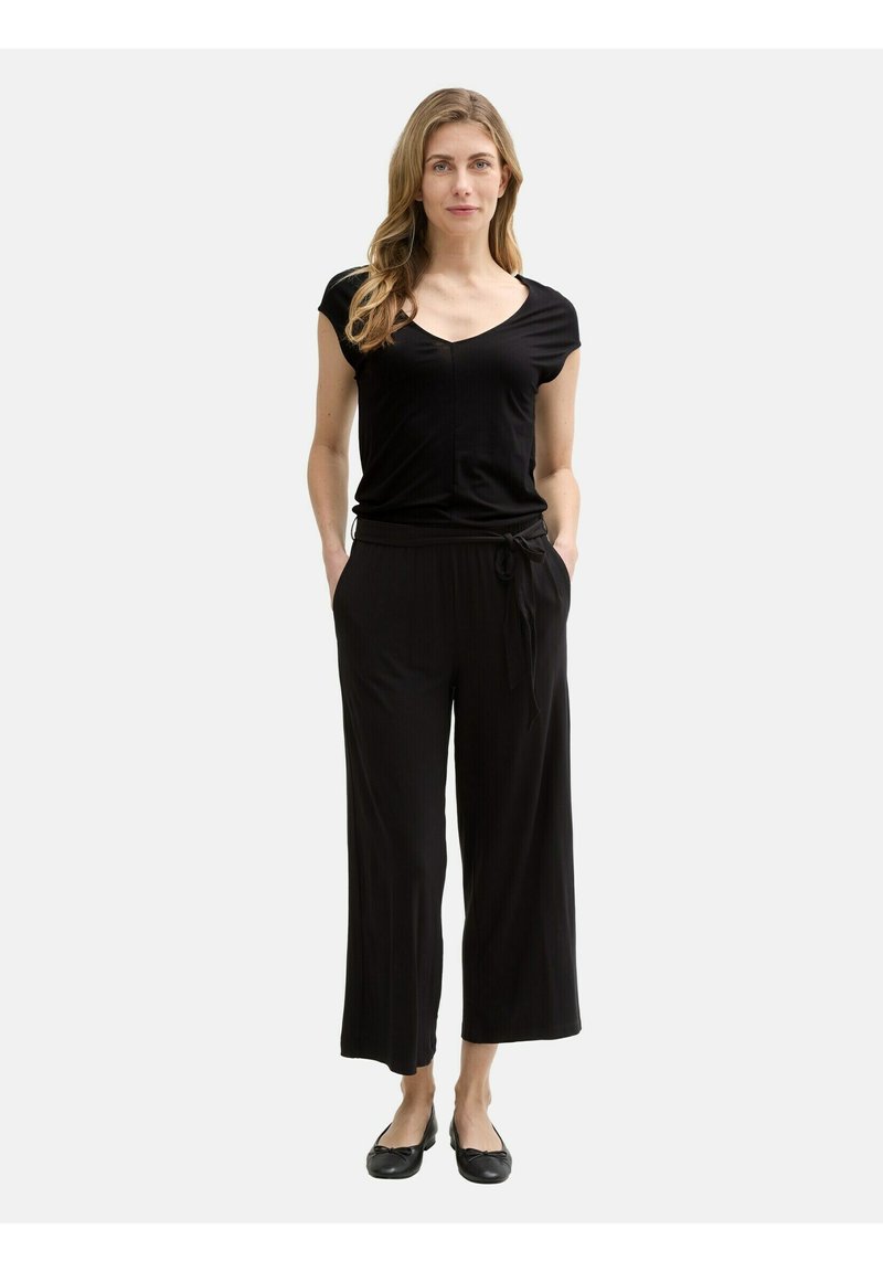 Tuta nera con maniche corte, scollo a V, vita cinched, pantaloni a gamba larga e texture morbida. Abbinato a scarpe flat nere.