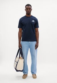 Jūrinės jėgos trumpomis rankovėmis marškiniai su priekinio logotipo dizainu, derinami su šviesiai mėlynomis džinsais. Modelis laiko kreminės spalvos rankinę su tamsiai mėlynais diržais.