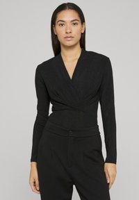 Body à manches longues noir avec un décolleté en V et un design devant croisé, doté d'une texture légèrement brillante. Associé à un pantalon noir ajusté.