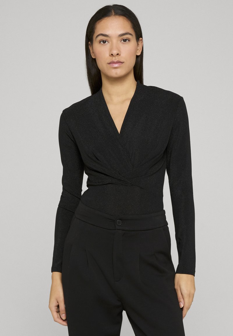 Body à manches longues noir avec un décolleté en V et un design devant croisé, doté d'une texture légèrement brillante. Associé à un pantalon noir ajusté.