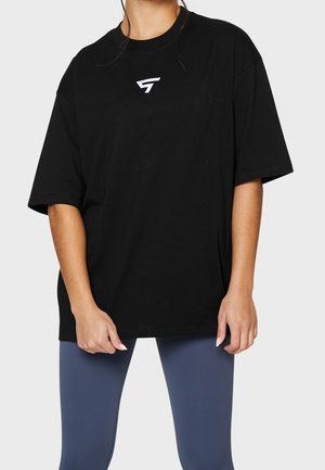 Woman wearing a loose black T-shirt with a small white logo on the chest and fitted blue leggings, standing against a plain background.