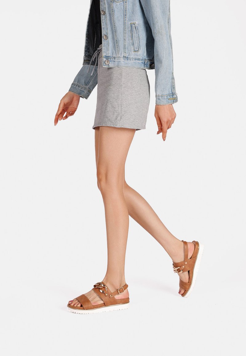 Sandali in pelle marrone con suola bianca, dotati di cinturini regolabili e dettagli in metallo. Abbinati a una gonna grigia e a una giacca di jeans.