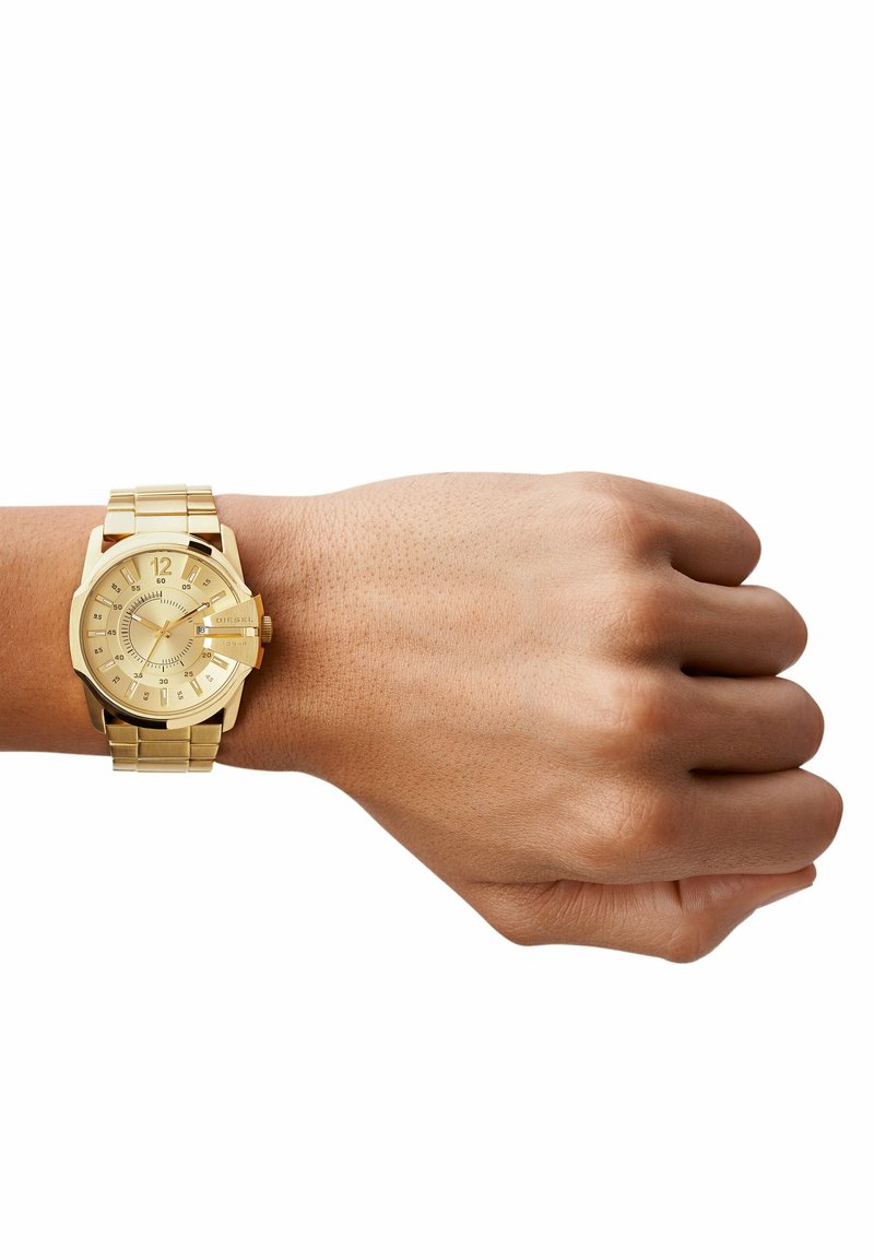 Orologio dorato con quadrante rotondo, indicatori chiari e bracciale metallico. Presenta una lunetta texturizzata e due sottoclassi. Indossato su un polso.