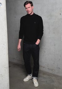 Homme en pull à col roulé noir, jean foncé et baskets blanches appuyé contre un mur en béton, une main dans la poche.