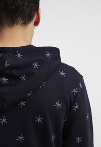 Námořnická mikina s kapucí, zdobená světle modrými vzory ve tvaru hvězdného výbuchu. Vyrobena z měkkého materiálu, s uvolněným střihem.