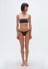 Γυναίκα που στέκεται ξυπόλητη, φορόντας καφέ σκούρο μπικίνι με λεπτές τιράντες, κοιτάζοντας προς τα αριστερά της σε απλό λευκό φόντο.