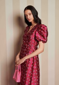 Vestido envelopado floral em fúcsia com mangas bufantes, apresentando um detalhe de laço na cintura. Segurando uma pequena bolsa de pérolas rosa.