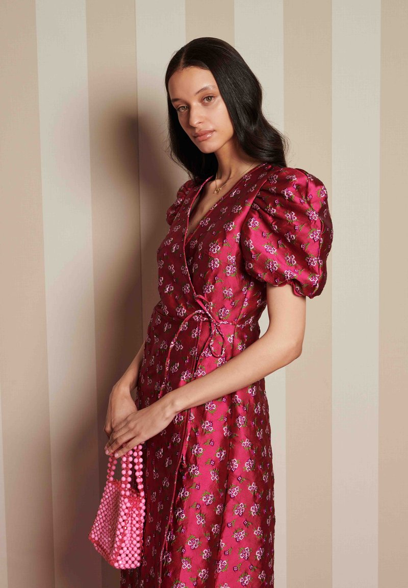 Vestido envelopado floral em fúcsia com mangas bufantes, apresentando um detalhe de laço na cintura. Segurando uma pequena bolsa de pérolas rosa.