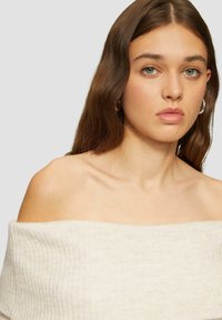 Off-shoulder geribbelde top in crème, met een zachte textuur en een brede halslijn. De stof is lichtgewicht en iets rekbaar.