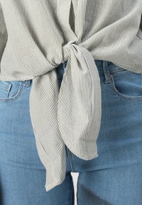 Striped blouse in light green and white, knotted at the waist. Worn with blue jeans, showcasing a casual and fitted design.