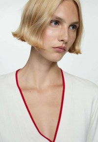 Top bianco con scollo a V profondo, con un bordo rosso a contrasto. La modella ha capelli biondi tagliati a casco e trucco minimale, con uno sfondo neutro.