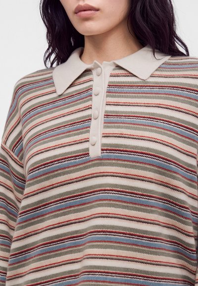 Femme portant un pull en maille à rayures horizontales beige, rouge, bleues et marron, avec un col beige et une patte de boutons.
