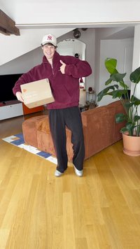 Gren tkaninski jopič bordo barve, črne trenirke, drži rjavo kartonsko škatlo z besedilom. Notranji prostor z zeleno rastlino in rjavo zofe.