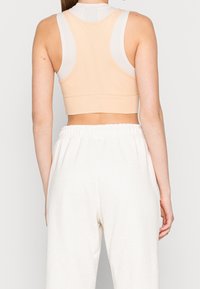 Kobieta nosząca beżowy i biały sportowy stanik z siateczki oraz luźne, jasnobeżowe spodnie z wysokim stanem, pokazana od tyłu na jednolitym tle.