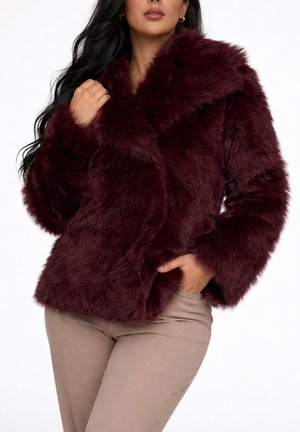 Vrouw draagt een bordeaux faux fur jas met een brede kraag, poseert met één hand nabij de kraag en de andere in de zak van beige broek.