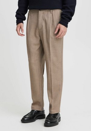 Mand iført beige skræddersyede bukser, sorte læder loafers og en mørk navystrikket sweater, stående mod en ensfarvet lys baggrund.