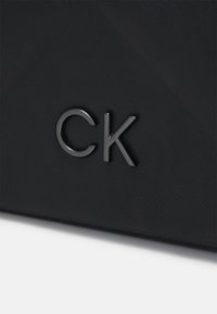 Čierna kožená taška s textúrovaným povrchom a kovovým logom CK v minimalistickej typografii. Ostrý dizajn s čistými líniami.