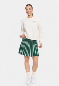 Sweatshirt crème avec un graphique vert et une jupe plissée à rayures vertes. Chaussures de sport blanches avec des chaussettes blanches et une tenue décontractée. Évite la texture.