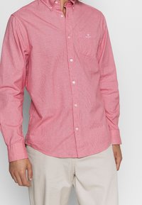 Rood-wit gestreept overhemd met lange mouwen en knopen, voorzien van een zak en een geborduurd logo op de borst, gecombineerd met lichtbeige broek.