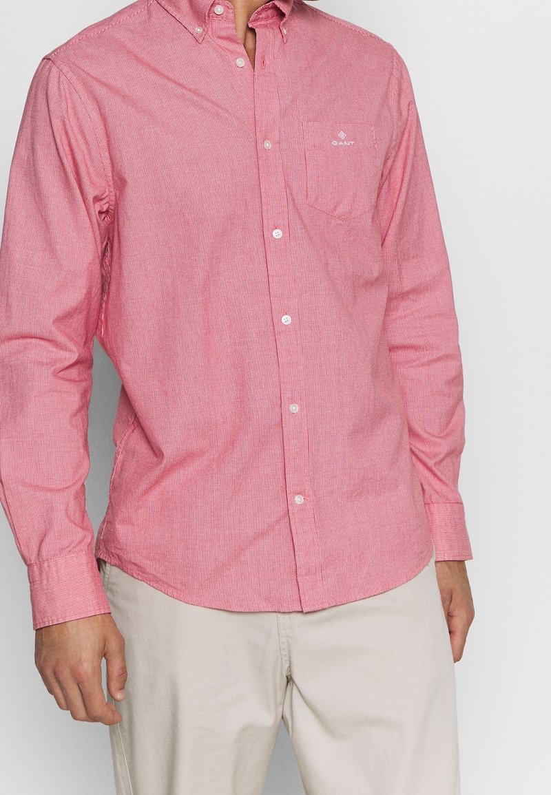 Rood-wit gestreept overhemd met lange mouwen en knopen, voorzien van een zak en een geborduurd logo op de borst, gecombineerd met lichtbeige broek.