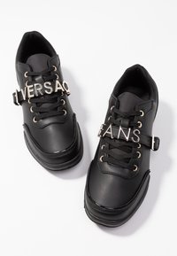 Baskets en cuir noir avec des accents en métal doré, présentant des lacets et des sangles affichant le texte "VERSA" et "JEANS". Texture lisse, bout arrondi.