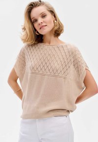 Beżowy sweter dzianinowy z diamentowym wzorem, szerokimi rękawami i luźnym krojem, zestawiony z białymi spodniami dla casualowego wyglądu.