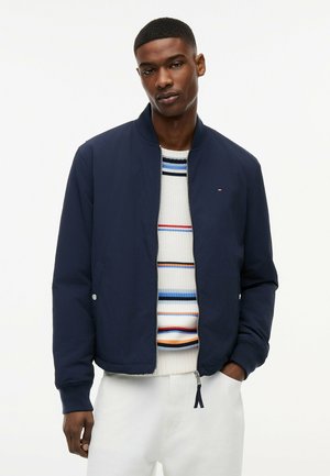 Młody mężczyzna w granatowej kurtce bomberce na białym swetrze w paski i białych spodniach, stojący z jedną ręką w kieszeni na białym tle.