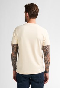 Svetlé béžové tričko s krátkým rukávem a kulatým výstřihem, vyrobené z jemné bavlny. Obsahuje viditelné tetování na obou pažích a tmavě modré kalhoty.