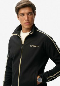 Chaqueta negra con cremallera y franjas doradas, de tela suave y cuello alto. Presenta un logo en el pecho y bolsillos laterales. Llevada con una camisa blanca debajo.