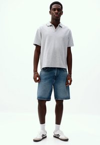 Polo shirt gri cu mânecă scurtă, pantaloni scurți din denim, șosete albe și adidași albi cu accente negre; croi lejer. Design minimalist cu linii curate.
