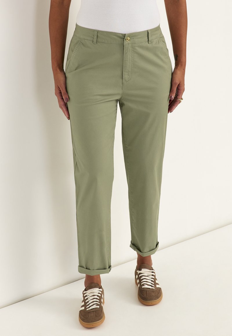 Persoon met licht olijfgroene opgerolde broek, wit shirt en bruine sneakers met witte veters tegen een effen achtergrond.