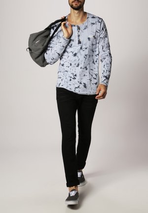 Chemise à manches longues bleu clair avec un motif tie-dye, col boutonné, jeans noirs et baskets grises, avec un sac à dos gris en bandoulière.