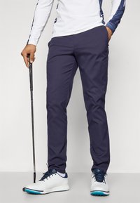 Navy golf pants, slim fit, made of lightweight fabric; matched with white and blue golf shoes. Person holds a golf club.