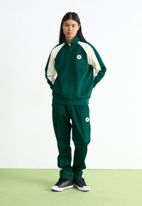 Πράσινο μπουφάν με φερμουάρ και κρεμ λεπτομέρειες, ταιριαστά πράσινα παντελόνια και μαύρα αθλητικά παπούτσια με λευκές σόλες και λογότυπο αστεριού στην αριστερή πλευρά.