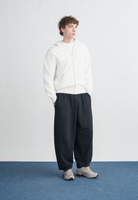 Camisola com capuz branca com fecho de correr e acabamento texturizado, combinada com calças de treino pretas largas. Acessórios incluem ténis cinza claro. Design simples e confortável.