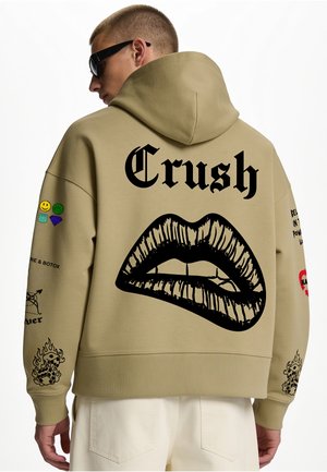 Mann trägt einen beigen Hoodie mit dem Wort "Crush" und einer großen schwarzen Lippen-Grafik auf dem Rücken sowie verschiedenen kleinen Patches an den Ärmeln.