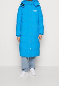 Ljusblå puffjacka med huva, med en logotyp, tryckknappar och två sidofickor. Buren över ljusblå jeans och vita sneakers.