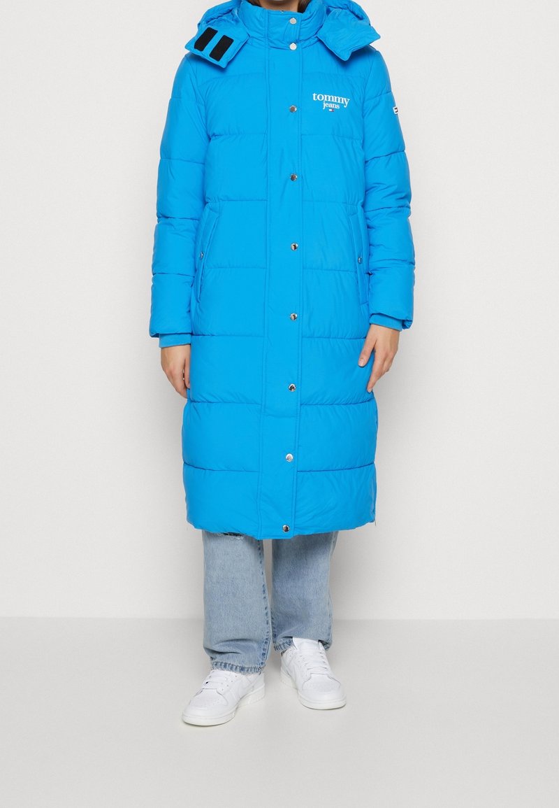 Ljusblå puffjacka med huva, med en logotyp, tryckknappar och två sidofickor. Buren över ljusblå jeans och vita sneakers.