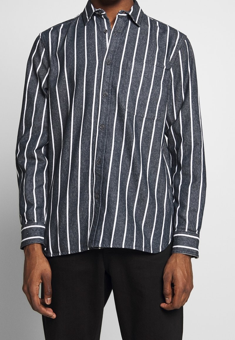 Camisa de botones de rayas en denim oscuro con rayas verticales blancas, que presenta un bolsillo de parche y mangas con puños. Textura suave, diseño casual.