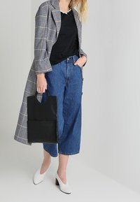 Lange geruite jas, zwarte gerimpelde top, blauwe wijde jeans, witte blokhak schoenen en een zwarte tas met een ronde handgreep.
