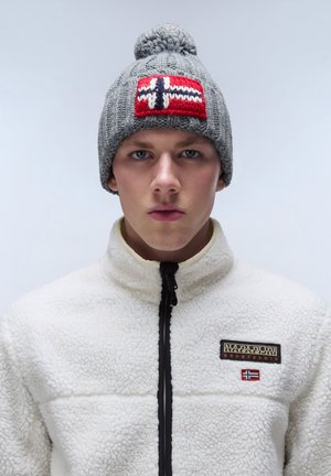 Gorro de punto gris con un parche de bandera noruega en rojo, blanco y azul. Textura suave y pompon en la parte superior. Usado con una chaqueta de forro polar blanca.